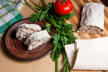 Two pieces of pita bread or lavash roll with cottage cheese or curd, chicken, tomatoes and herbs - dill, onion, parsley on brown clay plate..