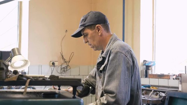 Mature caucasian man working in shabby car service, cylindrical boring of connecting rod bushing on old lathe. Wearing blue jeans jacket and cap. Shot with Sony a7s and Atomos Ninja Flame indoors.