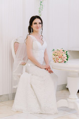 Beautiful bride perfect style. Wedding hairstyle make-up dress and bride's bouquet. Young attractive bride on chair in white room at studio