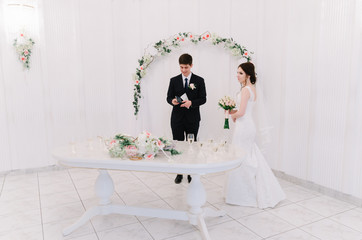 The bride in a white dress and groom in a blue suit are standing near an arch in the room and holding a wedding bouquet. Happy newlyweds