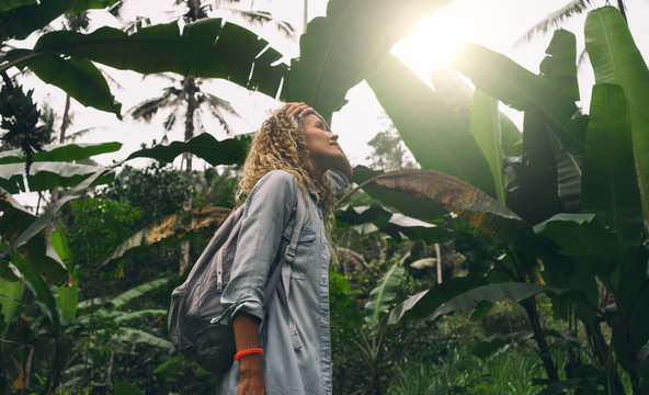 View from the bottom photo of a beautiful caucasian female with curly hair standing amid the huge green leaves and impressed with wild tropical nature during journey through the jungles.