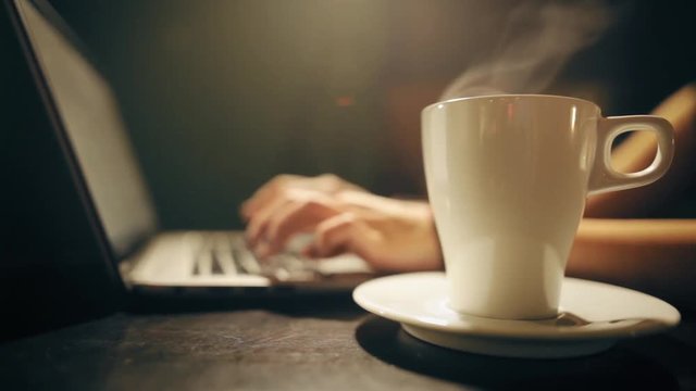 Still shot of a woman's hands typing on a laptop keyboard, and drinking a cup of hot coffee.