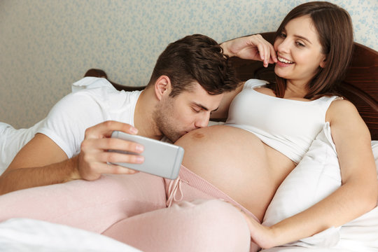Attractive young pregnant couple taking selfie while lying in bed
