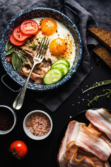 Breakfast set. Pan of fried eggs with bacon, fresh tomato, cucumber, sage and bread on dark serving board over black background, top view