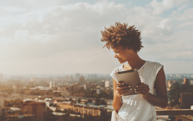 Cheerful young black curly female leaning on railing of balcony of skyscraper, holding digital tablet and looking down on city from high above; with copy space place for text, your message or advert - Powered by Adobe