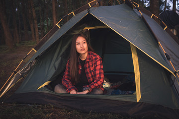 Asian woman looking out of the door of a tent in the woods with a red shirt. Trekking, camping, nature and outdoor concept.