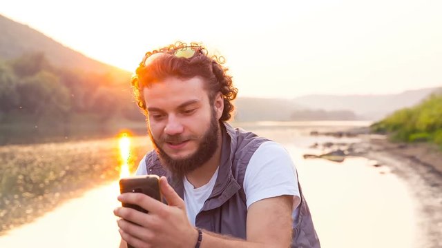 Young handsome man talking on a smartphone in the outdoors. The tourist speaks on Skype sitting on the riverbank.