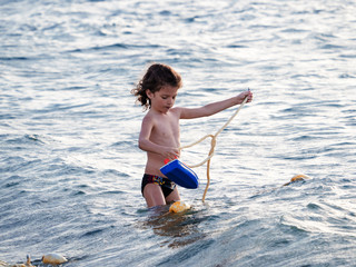 A little boy is playing with a toy boat, standing in the ocean.