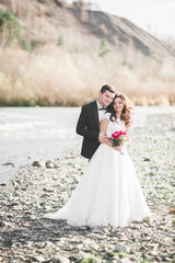 Fototapeta na wymiar Elegant stylish happy brunette bride and gorgeous groom on the background of a beautiful river in the mountains