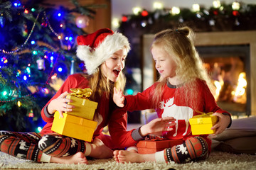 Happy little sisters wearing Christmas pajamas playing by a fireplace in a cozy dark living room on Christmas eve