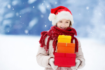 Adorable little girl wearing Santa hat holding a pile of Christmas gifts on beautiful winter day