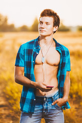 a handsome young man with muscular torso listening to music in white headphones and holding a smartphone in his hand. He is dressed in a shiny blue shirt and jeans shorts, is in a field outside city