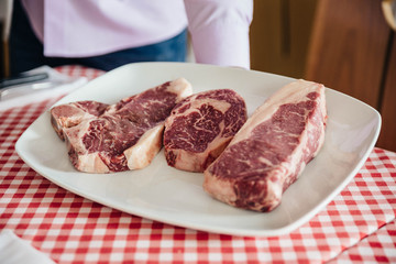 Raw wagyu beef cuts from left to right: T-Bone Steak, Rib Eye and Boneless Top Loin Steak in white plate on red and white pattern tablecloth.