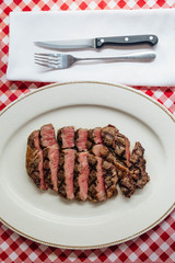 Top view of Sliced medium rare charcoal grilled wagyu Ribeye steak in white plate on red and white pattern tablecloth with cutlery.