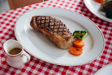 Charcoal grilled boneless wagyu Top Loin steak served with BBQ sauce and baked potato in white plate on red and white pattern tablecloth.