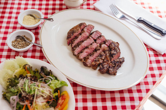 Sliced medium rare charcoal grilled wagyu Ribeye steak in white plate on red and white pattern tablecloth with salad and cutlery.