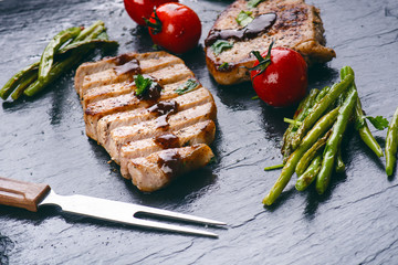 Grilled pork chops, steaks with vegetables, tomatoes, beans and sauce on a black slate. Fresh meat with foam. Dark background. Grilled and barbecue concept. 