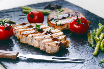 Grilled pork chops, steaks with vegetables, tomatoes, beans and sauce on a black slate. Fresh meat with foam. Dark background. Grilled and barbecue concept. 