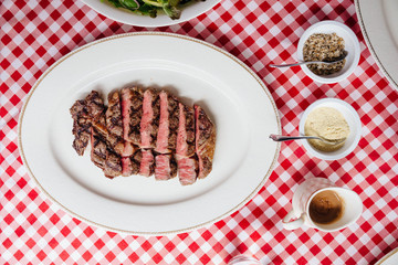 Top view of Sliced medium rare charcoal grilled wagyu Ribeye steak in white plate on red and white pattern tablecloth with seasoning and BBQ sauce.
