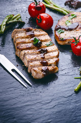 Grilled pork chops, steaks with vegetables, tomatoes, beans and sauce on a black slate. Fresh meat with foam. Dark background. Grilled and barbecue concept. 