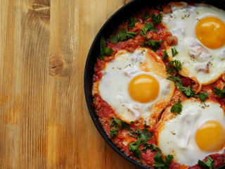 A pan of fried eggs with tomato sauce and parsley on a wooden background. Shakshuka a traditional meal of the Jewish cuisine