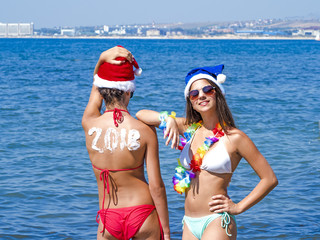 Two girlfriends in a Santa hat during a beach holiday. Girl with the inscription 2018 on the back.  Happy new year