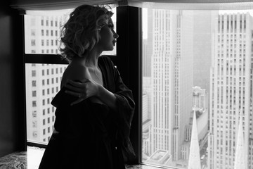 Sexy woman in black silk robe stands with naked shoulders before a panoramic window in New York