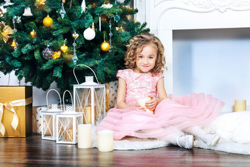 A sweet blonde girl with curls in a pink dress sits in the New Year decorations at the white...