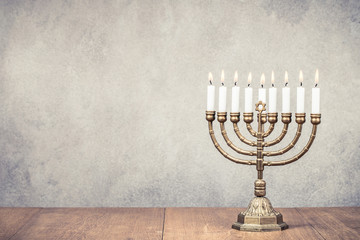 Bronze Hanukkah menorah with burning candles on wooden table front old vintage concrete wall background. Holiday greeting card concept. Retro instagram style filtered photo - obrazy, fototapety, plakaty