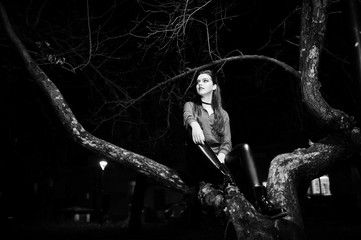 Black girl in red shirt and bright make-up on night street sitting on scary tree outdoor. Halloween theme.