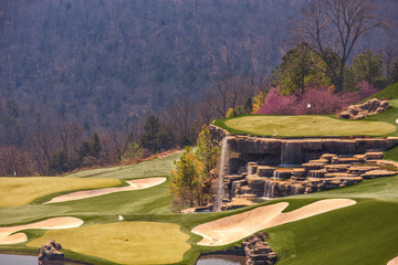 A golf course with sand bunkers and a waterfall in a valley surrounded by a forest of bare trees in...