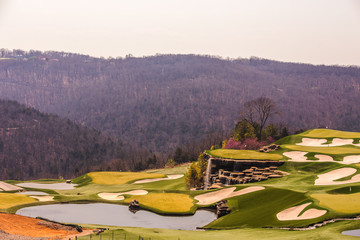 A golf course with sand bunkers and a waterfall in a valley surrounded by a forest of bare trees in springtime