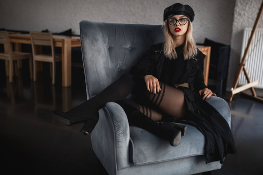 Young sexy woman in provocative pose sitting on the arm chair. Fashion and trendy clothes. Black leather cap, coat and shoes. Outumn stlyle. Inrerior photo.