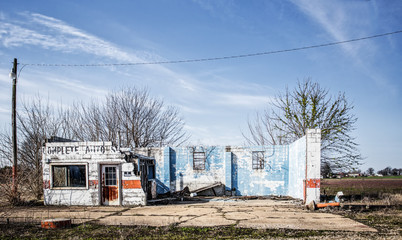 An old crumbled and faded brick auto service center with front wall gone and cement pad in front under blue wispy sky