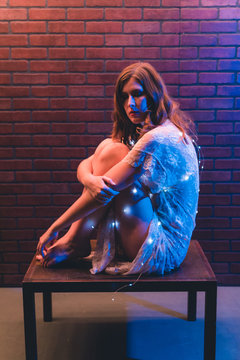 Young caucasian woman in sheer dress poses with string lights against a brick wall