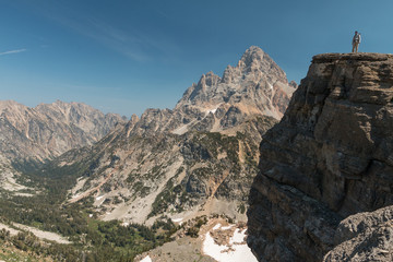 A hiker stands on a cliff in front of Grand Teton