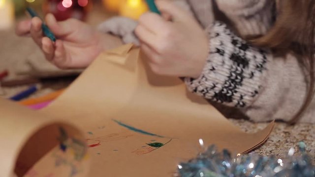A pretty little girl draws a drawing for Santa Claus near a beautifully decorated Christmas tree. Close-up. Handles of the little girl draw a picture