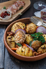 Bowl of oven roasted hassleback Potato with garlic, carrots and onion on rustic black wooden surface