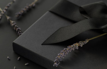 Black gift box on a dark contrasted background, decorated with a textured bow and lavender,...