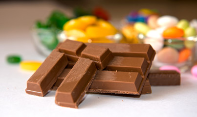 A stack of milk chocolate bars with broken pieces on a white surface. In the background, there are glass bowls filled with colorful candies, including yellow sweets and green treats