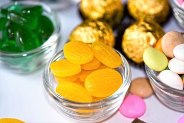 A glass bowl filled with bright yellow hard candies, surrounded by other sweet treats including green jelly candies, golden-wrapped chocolates, and colorful sugar-coated candies on a white surface
