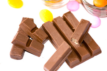 A close-up of milk chocolate bars broken into pieces, revealing a crispy wafer inside. The rich chocolate is accompanied by colorful candies, creating a tempting and indulgent dessert display