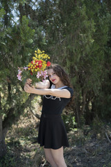 Pretty young lady in short black dress with white lapel holding and show a bouquet of flowers, outdoor shot with trees in background