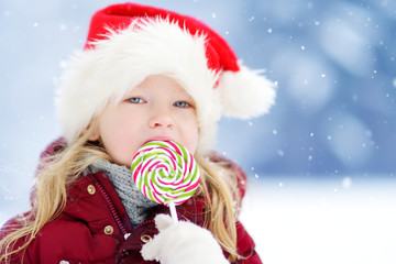 Adorable little girl wearing Santa hat having huge striped Christmas lollipop on beautiful winter day