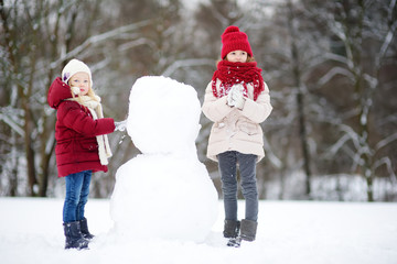 Two adorable little girls building a snowman together in beautiful winter park. Cute sisters playing in a snow.