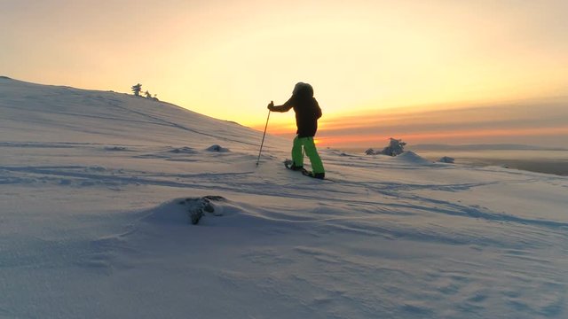 AERIAL Active woman in winter clothes snowshoeing on snowy mountain slope at sunrise. Young woman snowshoe hiking at winter sunset. Traveler snowshoe exploring snowy Lapland mountains in sunny morning