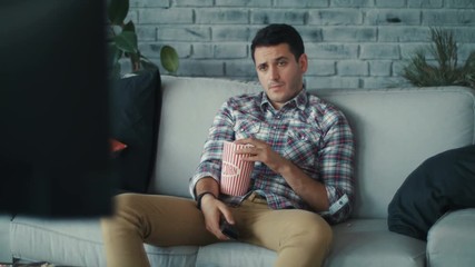 Close up young man watching the TV siting on the sofa with remote in his hands use phone in home handsome laziness looking television men shot on RED EPIC camera. Slow Motion Shot.