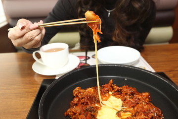 Korea Food in Restaurant on wooden table, stir fried spicy pork with cheese on brass plate, girl fun eating sticky cheese, focus food blur woman