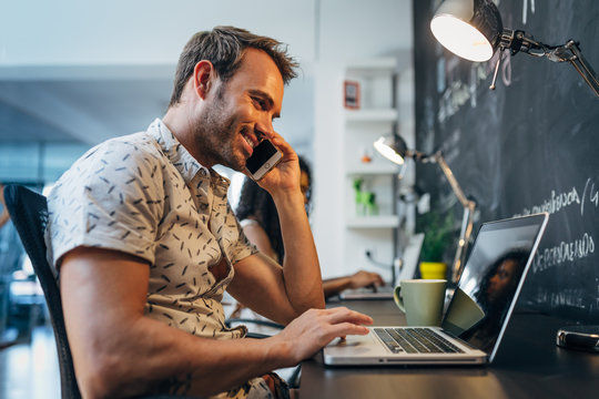 Happy Man Working on Laptop at the Office while Talking on Phone