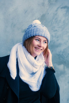 A beautiful young woman with dark hair in a large knitted white bactus scarf made of natural wool and black classic сoat  smiling and posing on the background of a stone gray wall